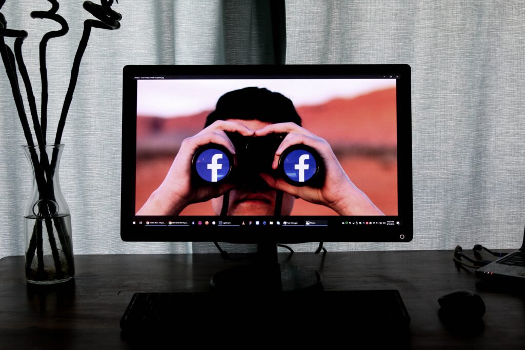 Desktop monitor with background image of a man looking through binoculars with the Facebook logo reflected in them.
